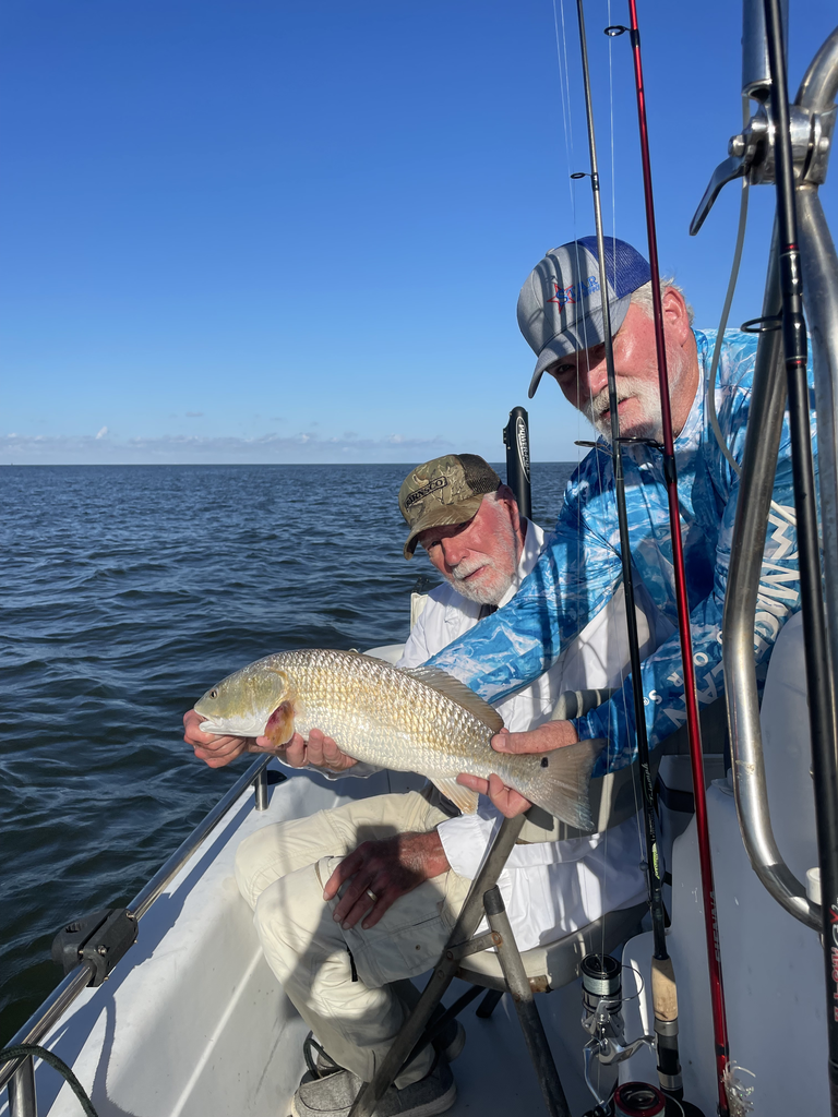Two men on a boat, holding a large fish, blue sky, and calm water in the background.