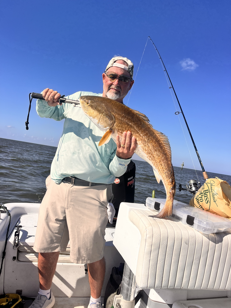 Man on a boat holds up a large redfish. Blue sky, water, and fishing rods are visible.