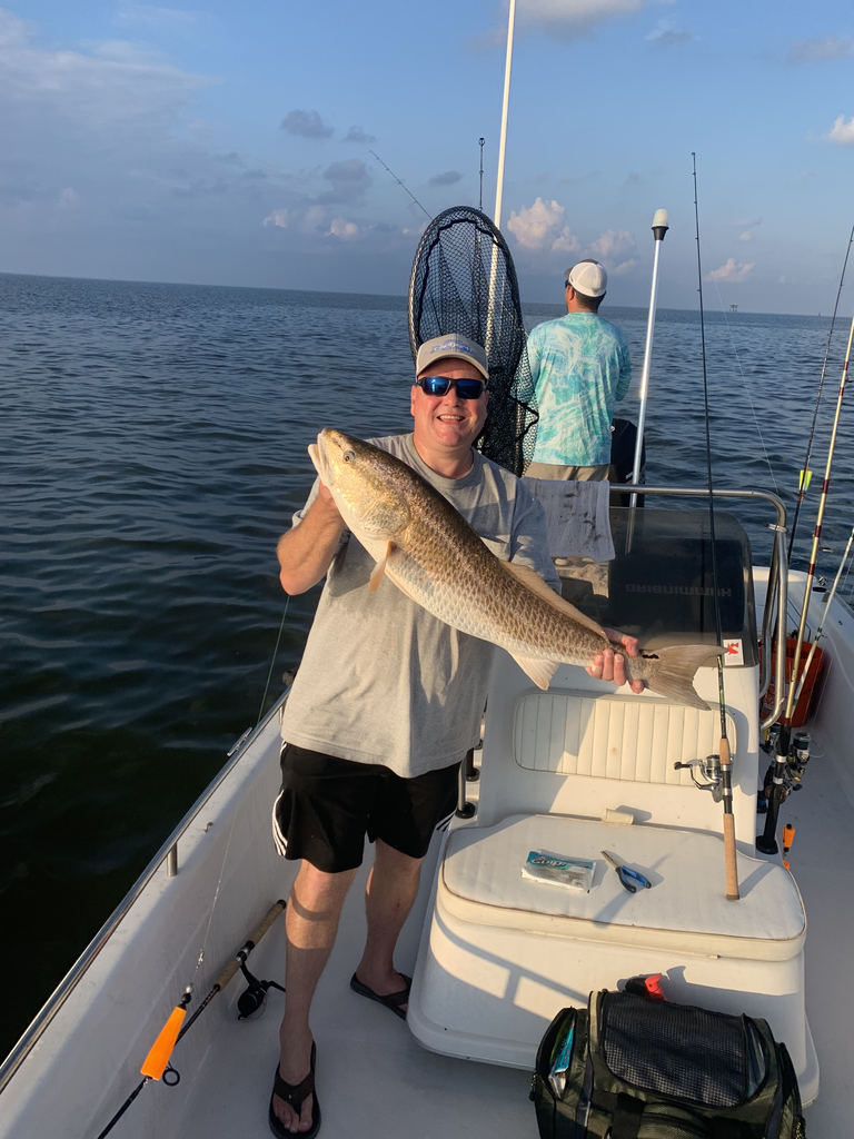 Man on boat holds a large fish, smiling. Another person stands in the background, rods visible, calm water.