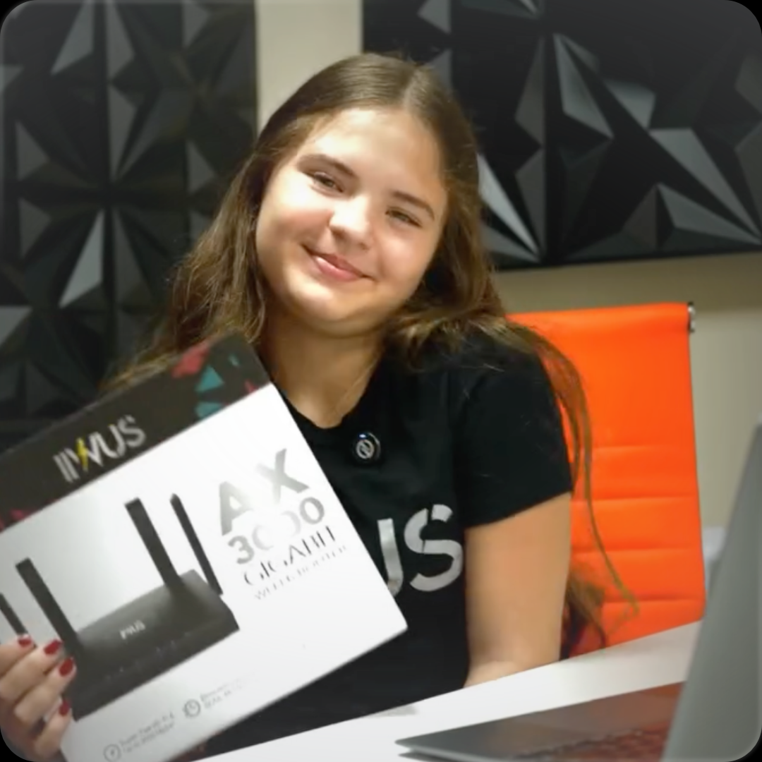 Smiling person holding a certificate, seated at a table with an orange chair and dark geometric wall behind.