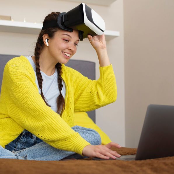 Woman using a VR headset beside a laptop, smiling on a couch in a bright room