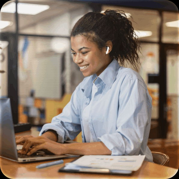 Woman typing on a laptop at a desk in a bright office, smiling while working.
