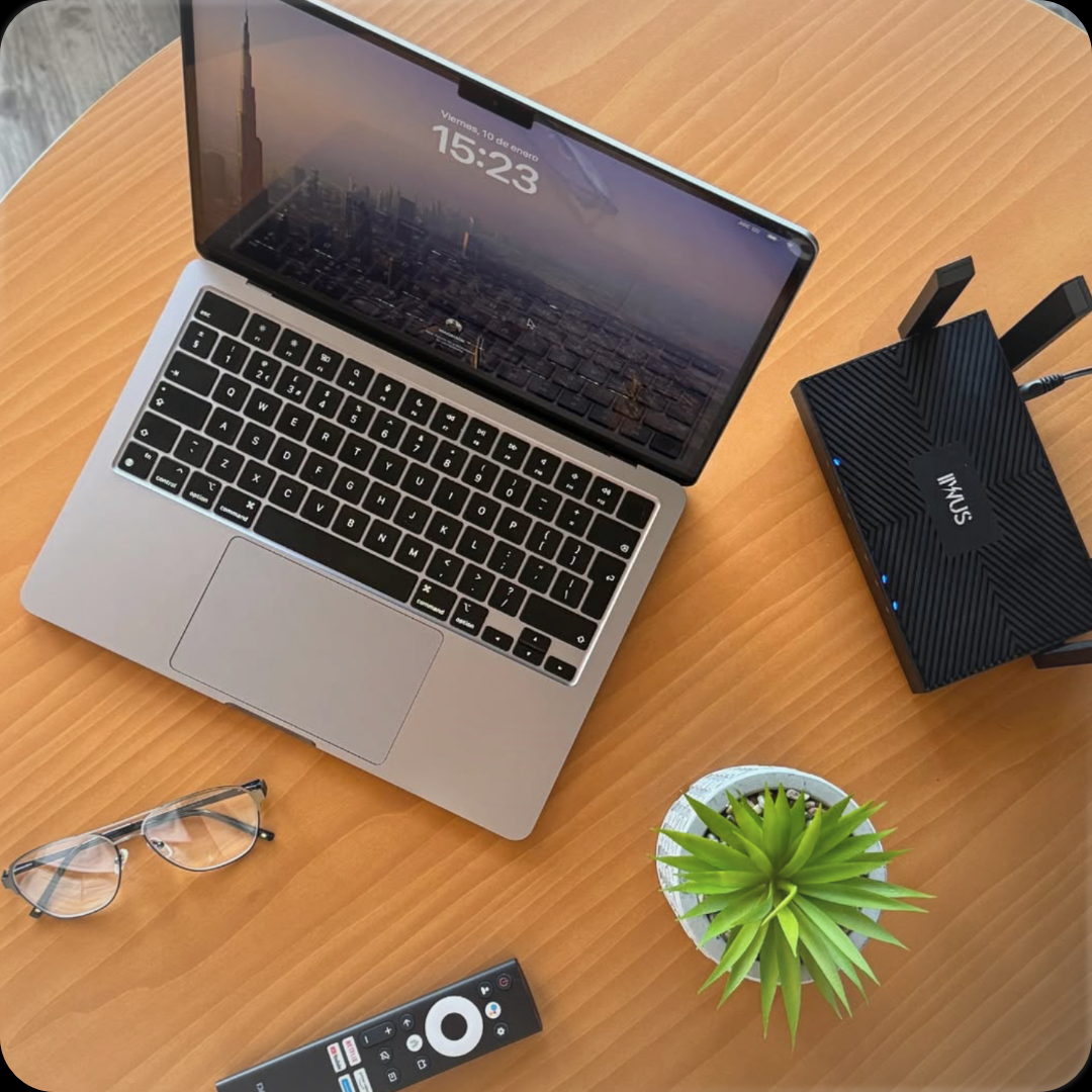Desk with open laptop, glasses, remote, small plant, and black device on a wooden tabletop.