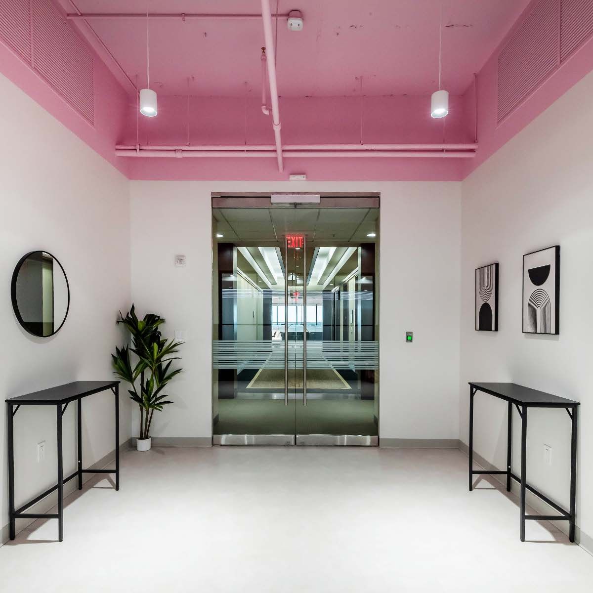 A modern office entrance with pink ceiling, glass doors, black console tables, and artwork on white walls.