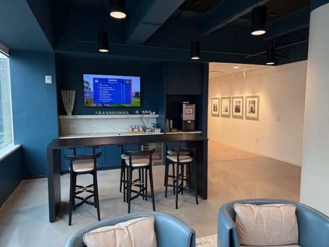 A modern lounge with a TV, bar, and seating. Dark blue walls and a neutral floor.
