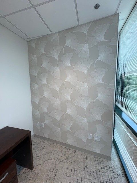 Office corner with gray patterned wallpaper, window with blinds, and a dark wooden desk.