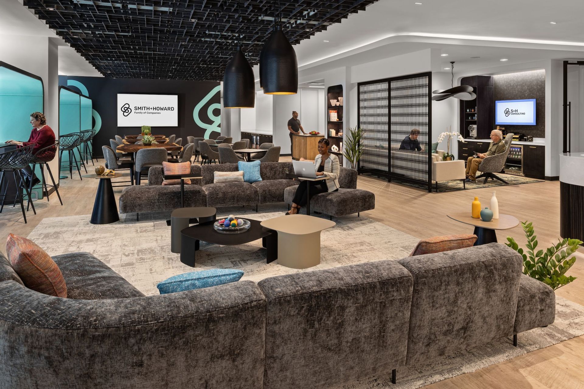 Modern office lounge with seating areas, tables, and people working. Dark ceiling and neutral color palette.