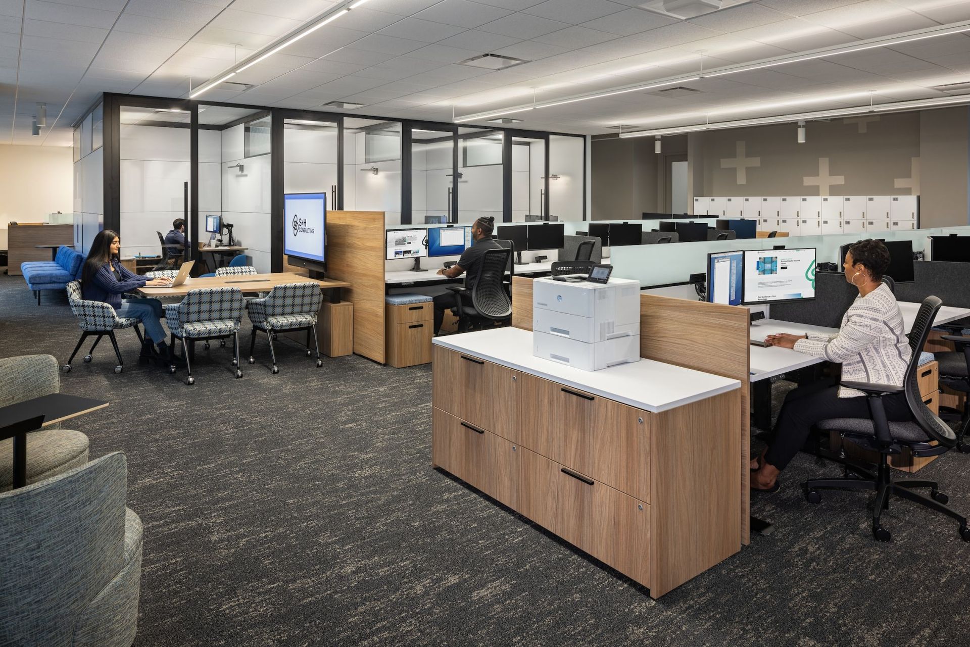 An open office with employees working at computers. Some areas have low dividers, wood and white desks, gray carpet.