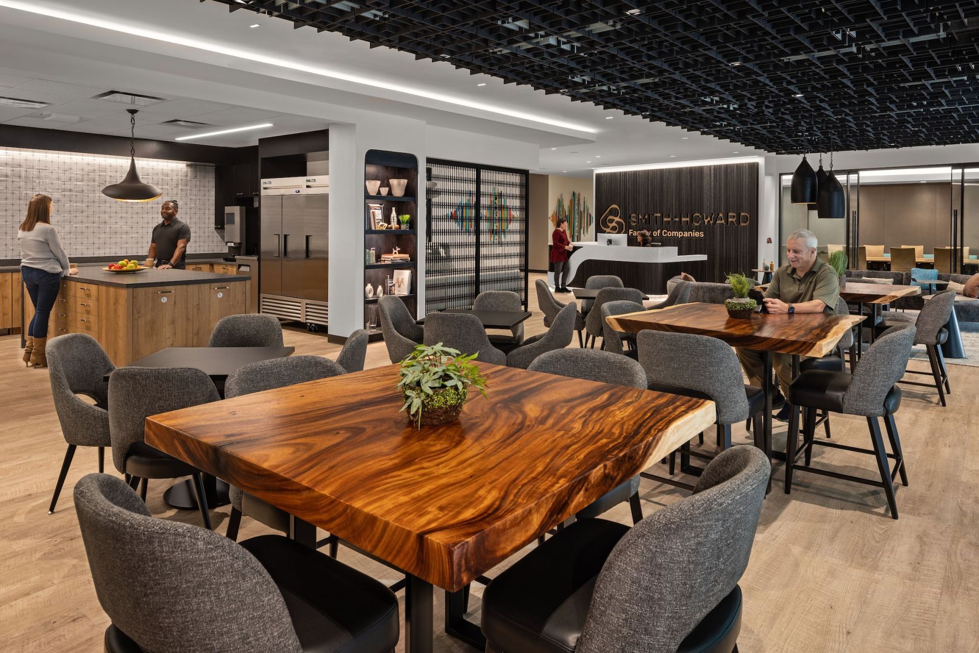 Modern office kitchen/dining area with wood tables, gray chairs, and people preparing food.