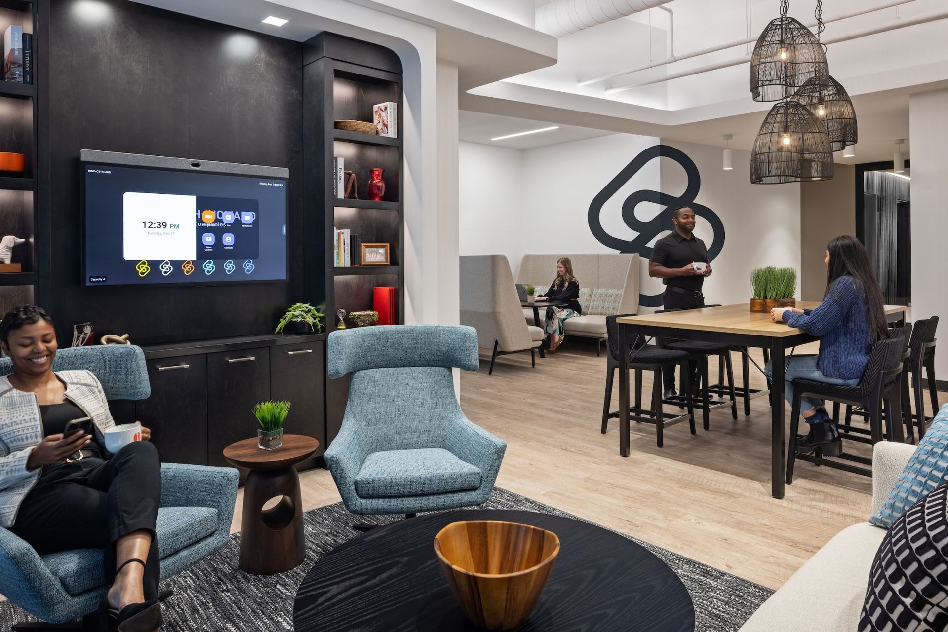 Cozy office lounge with seating areas. People sit at tables, chairs, and couches. A large screen displays content.