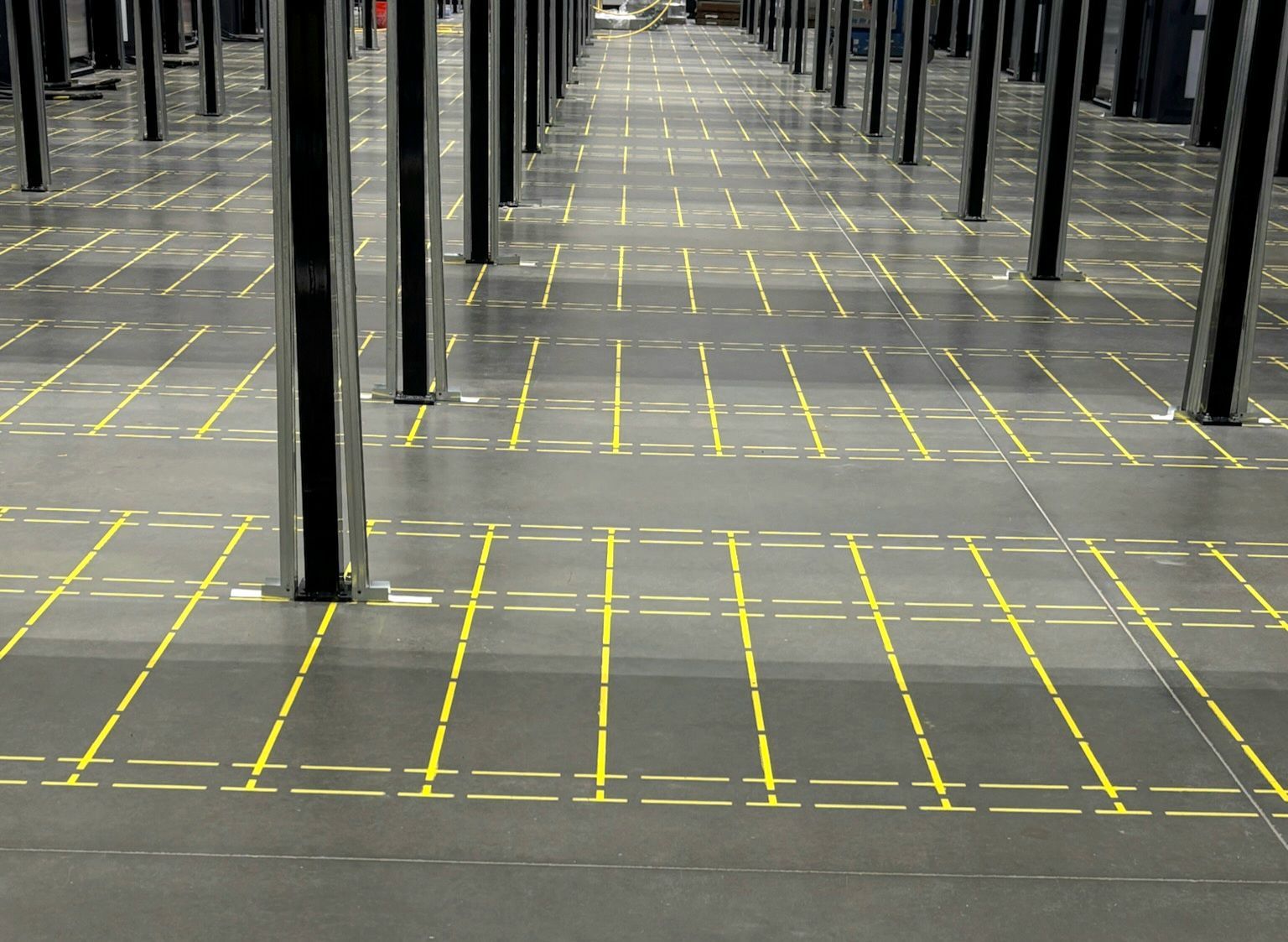 Rows of black support columns in a warehouse with yellow grid lines on the dark floor.