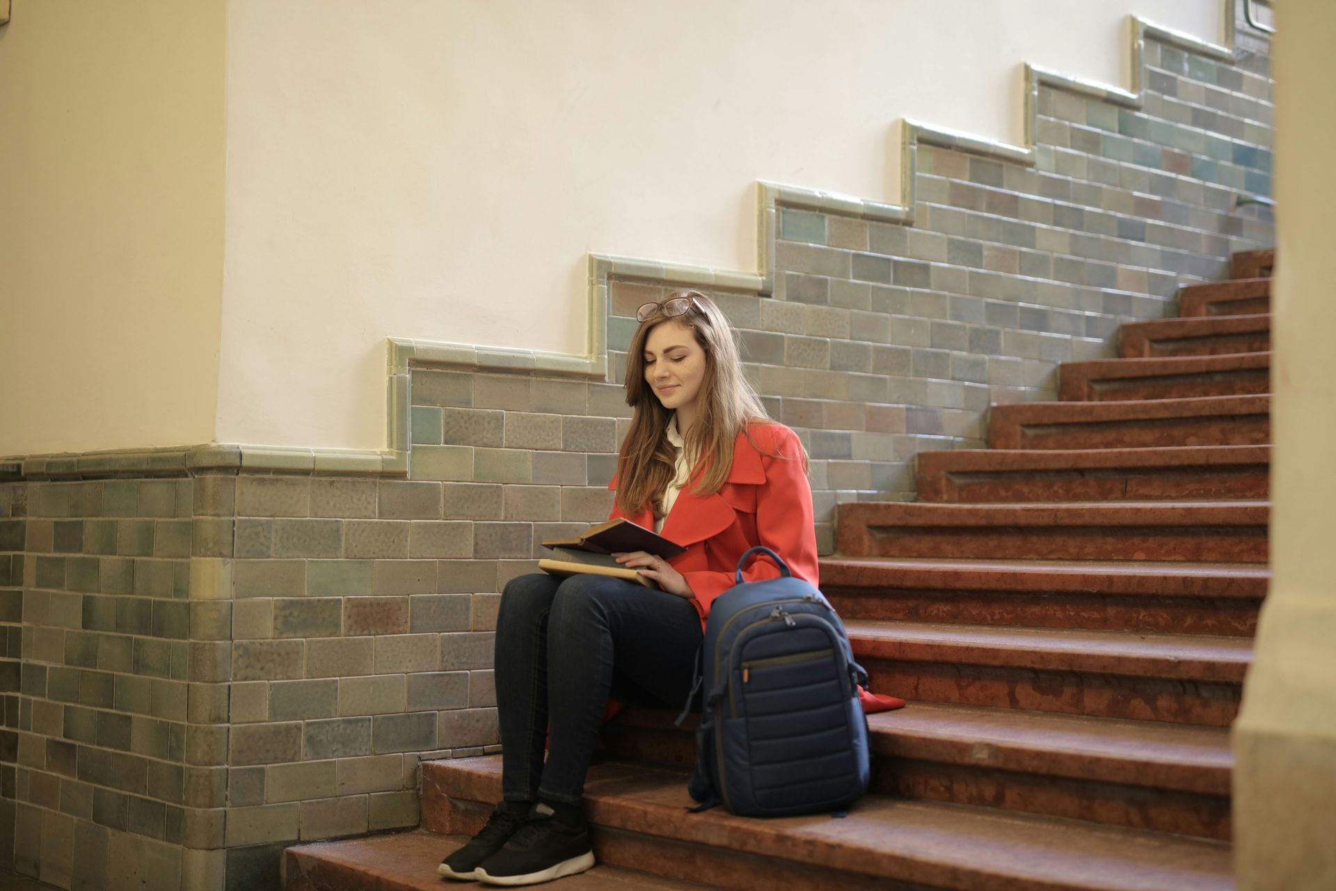 Woman in red jacket sits on stairs, reading a book. A backpack rests beside her.