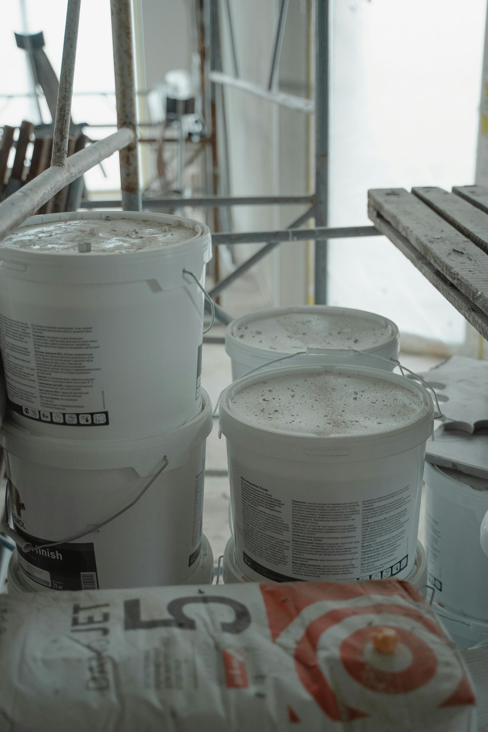 White buckets of material stacked on a construction site with scaffolding and a bag of building material.
