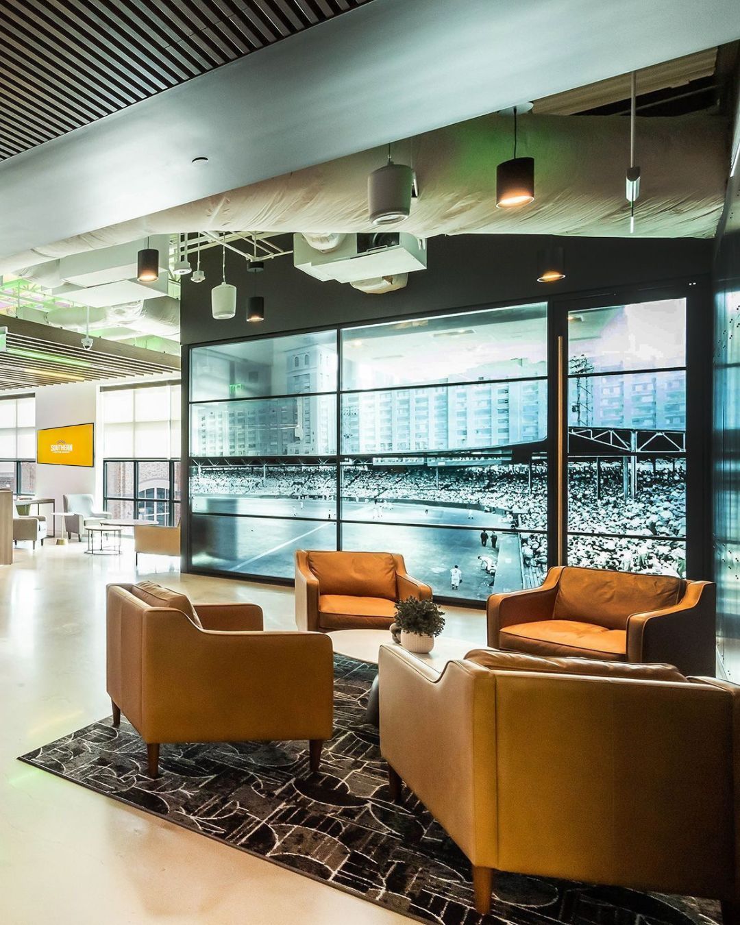 Lounge area with orange armchairs around a rug, facing a large screen displaying a sports stadium.