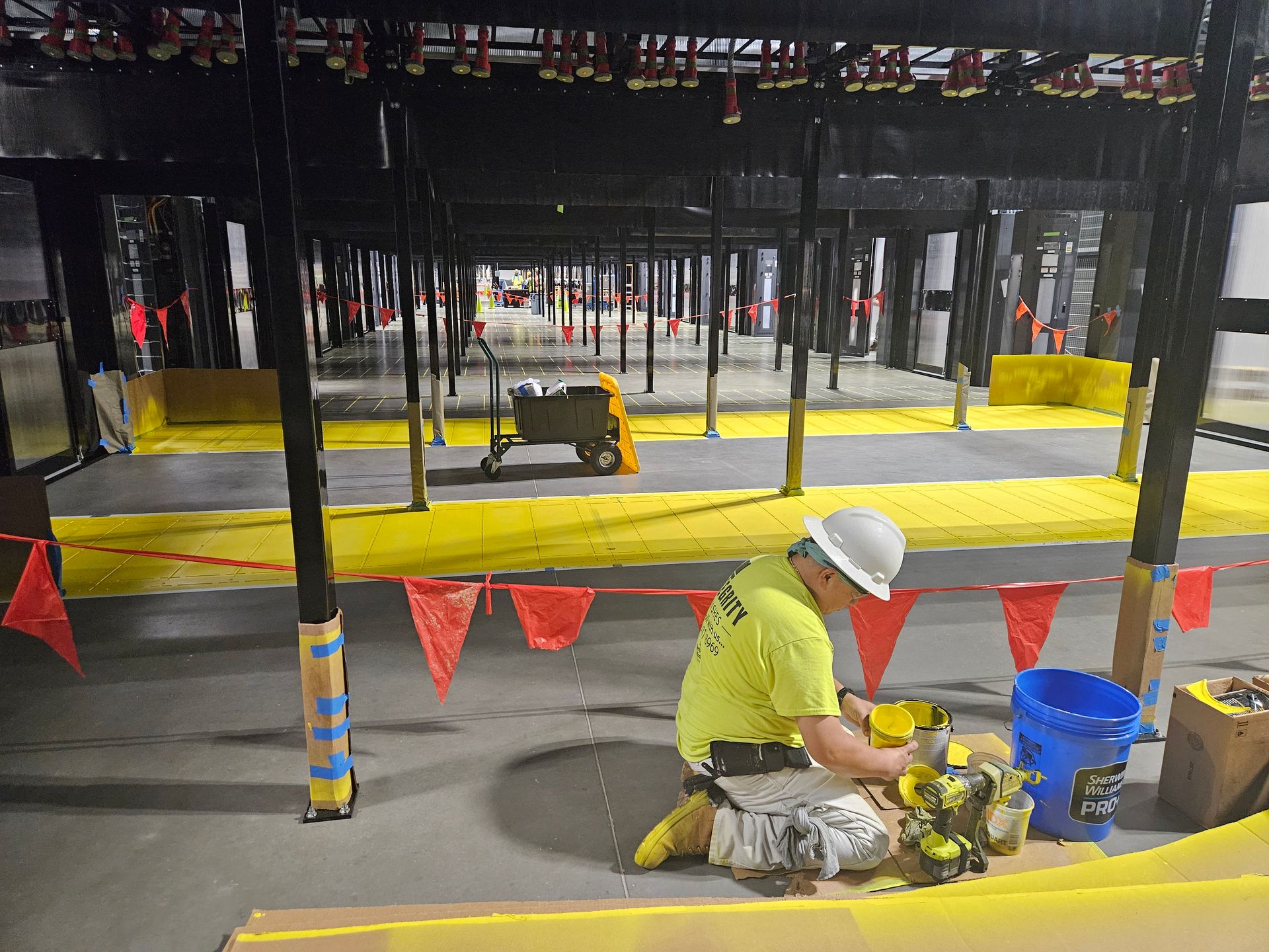 Construction worker kneels on floor, working. Yellow tape and safety cones surround workspace. Black beams overhead.