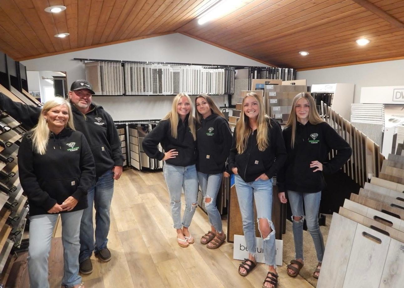 People standing inside a flooring showroom. They wear black hoodies and jeans. Wood flooring and samples surround them.