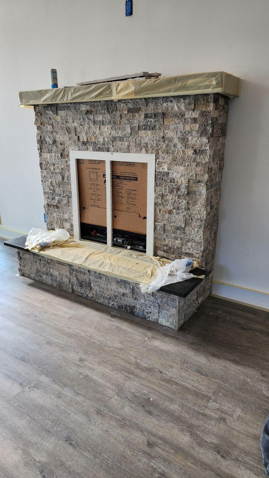 Stone-veneer fireplace under construction; grey and beige tones, white-framed opening, dark hearth, light brown flooring.