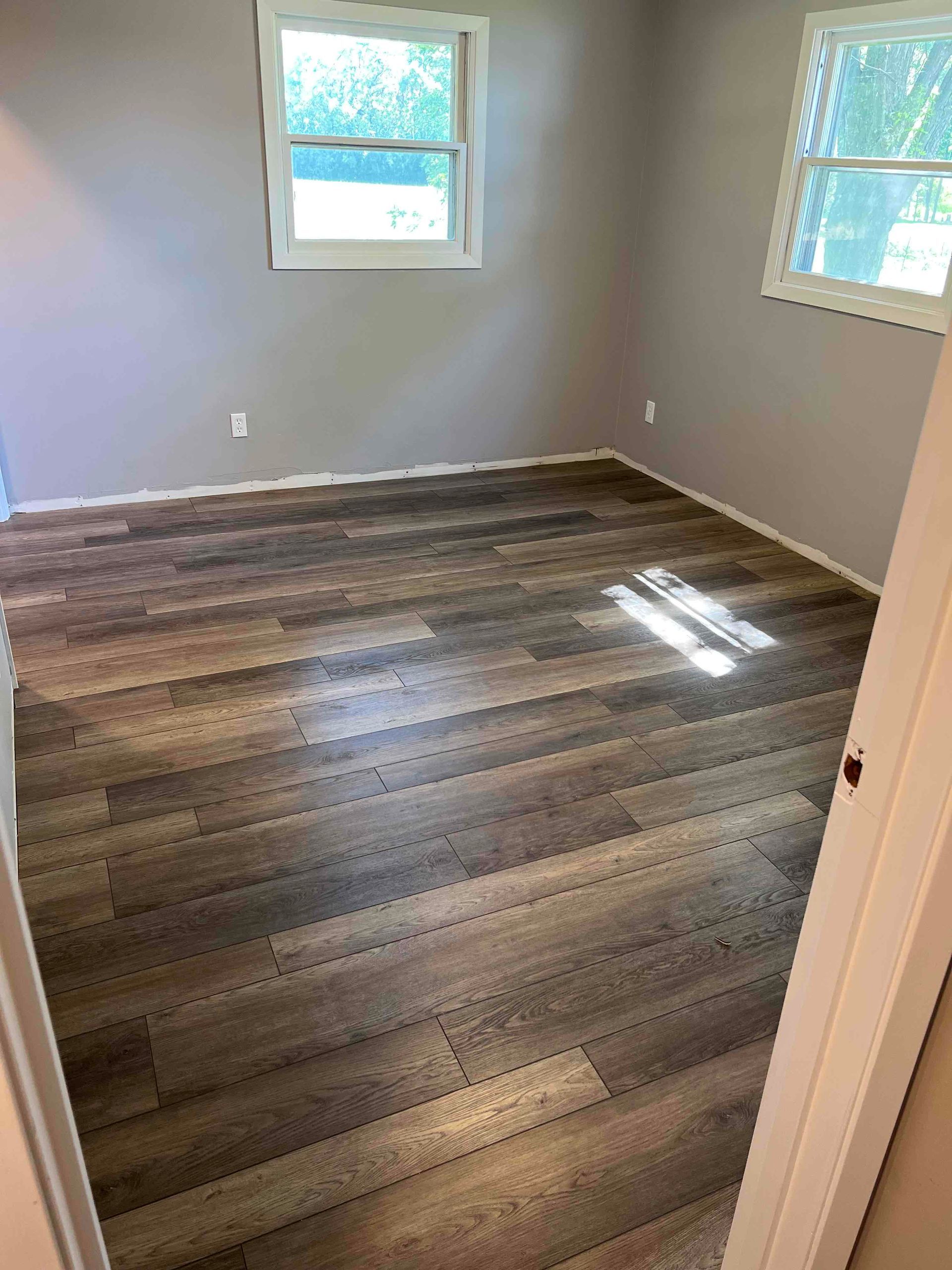 Empty bedroom with wood-look flooring, two windows, and light gray walls.