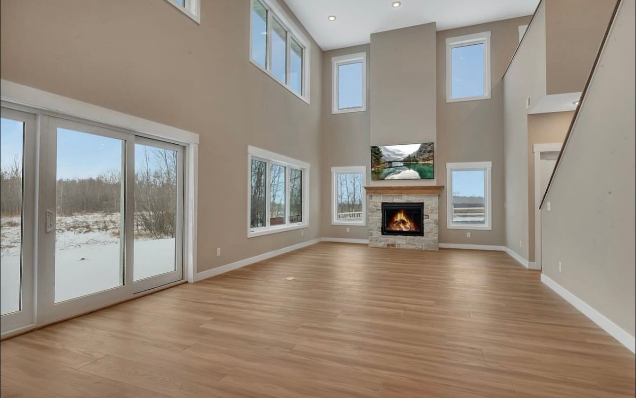 Empty living room with wood floor, fireplace, and large windows looking out to a snowy landscape.