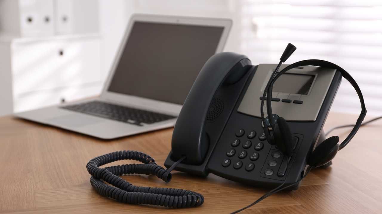 A telephone with a headset is sitting on a desk next to a laptop.