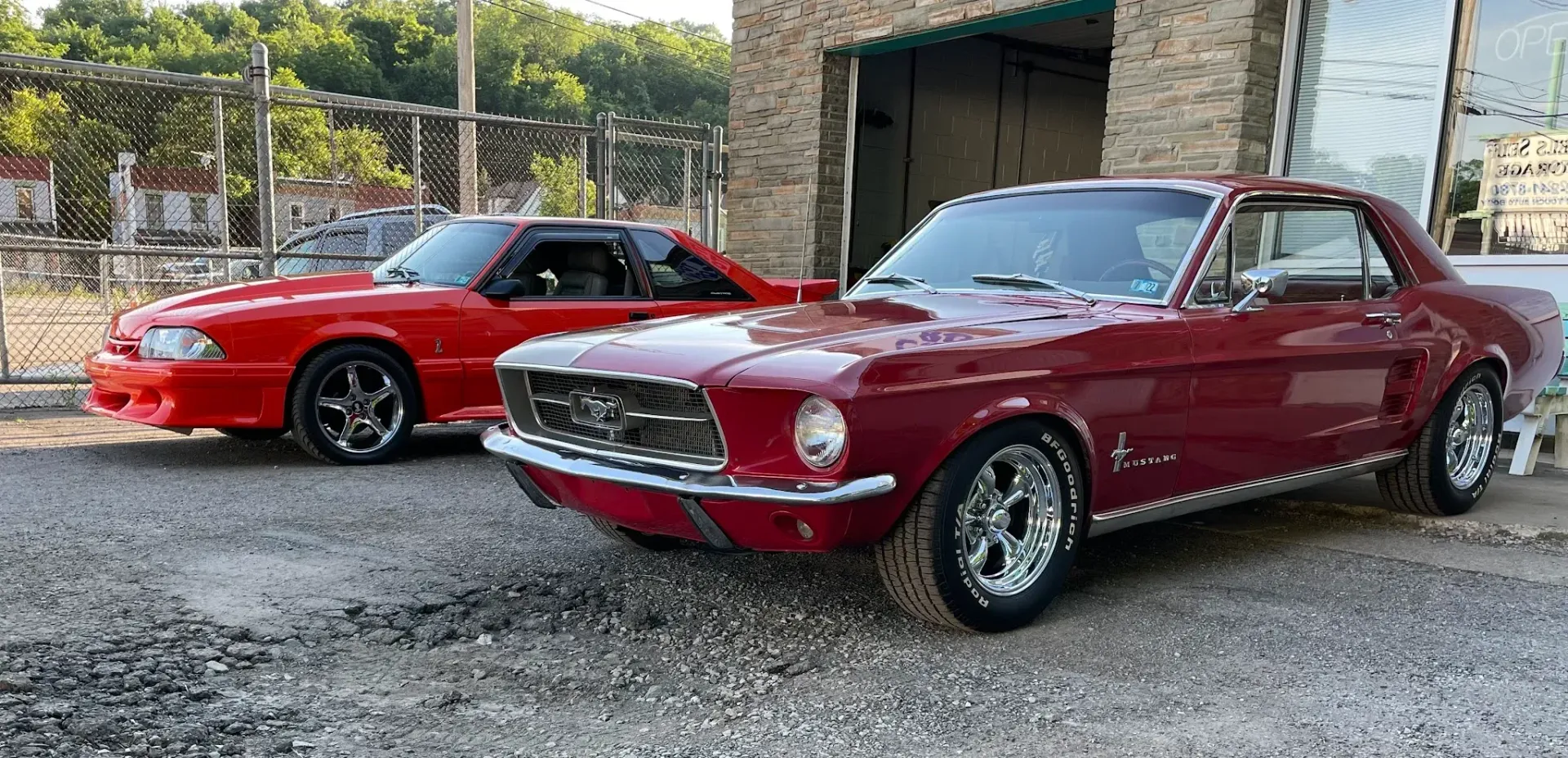 Two red classic cars parked on gravel outside a garage