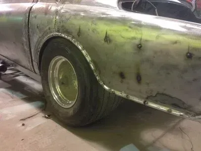 Close-up of a silver car rear quarter panel and tire in a garage, with visible dents and rust spots