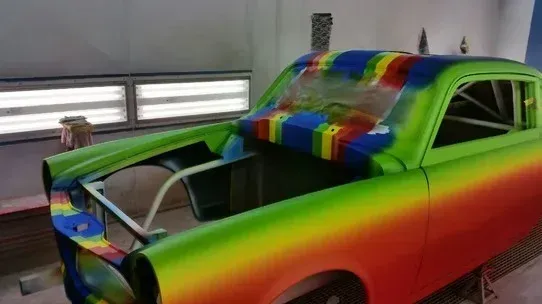 Rainbow-painted vintage car body in a garage, with hood open and no front grille or bumper.