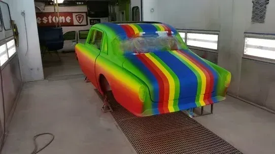 Rainbow-painted car in a garage booth, covered in bright stripes of spray paint.
