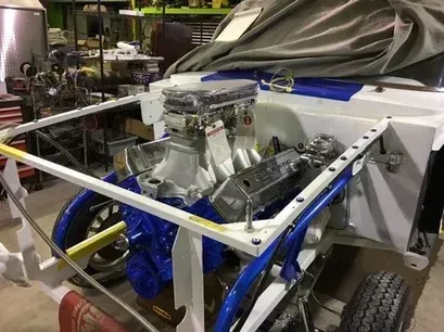 Mechanical chassis with exposed engine and large wheels in a workshop, partially covered by a tarp