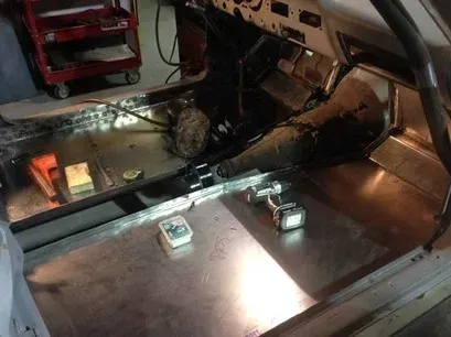 Car trunk interior with a metal floor panel, wiring, tools, and a wheel visible in the back.