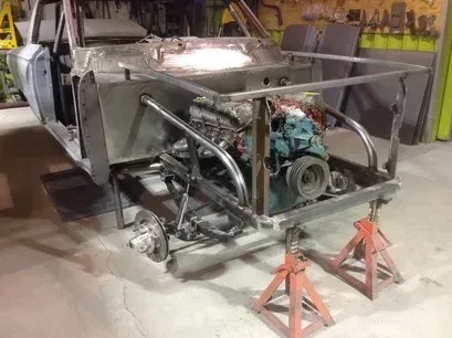Partially assembled silver car chassis on jack stands in a garage, with exposed engine and rear frame.