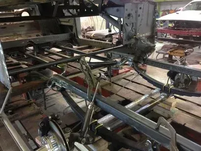 Close-up of a metal car chassis frame in an industrial workshop, with pipes and structural beams visible.