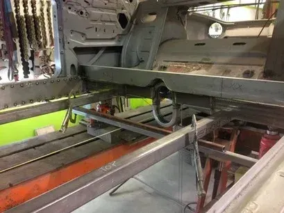 Interior of a stripped car frame with exposed metal beams and a ratchet strap across the floor.