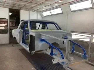 Classic car body in a spray booth, partially restored with exposed frame and blue masking tape.