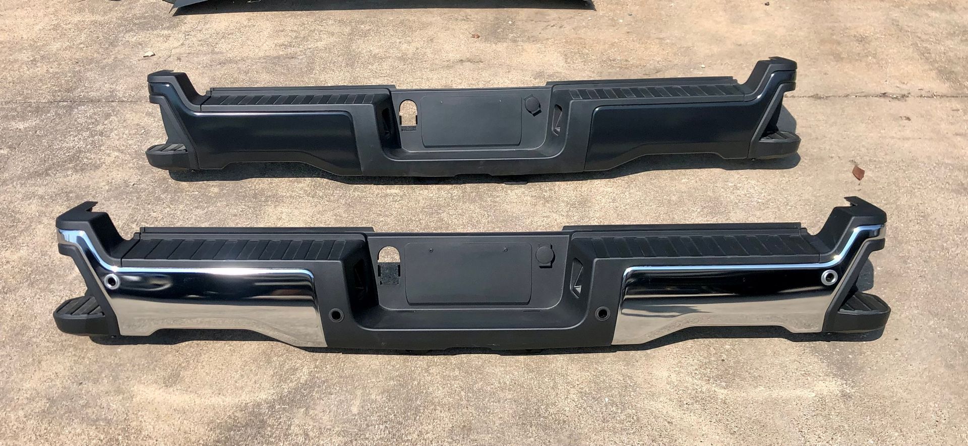 Two black and chrome bumpers are sitting on the ground.