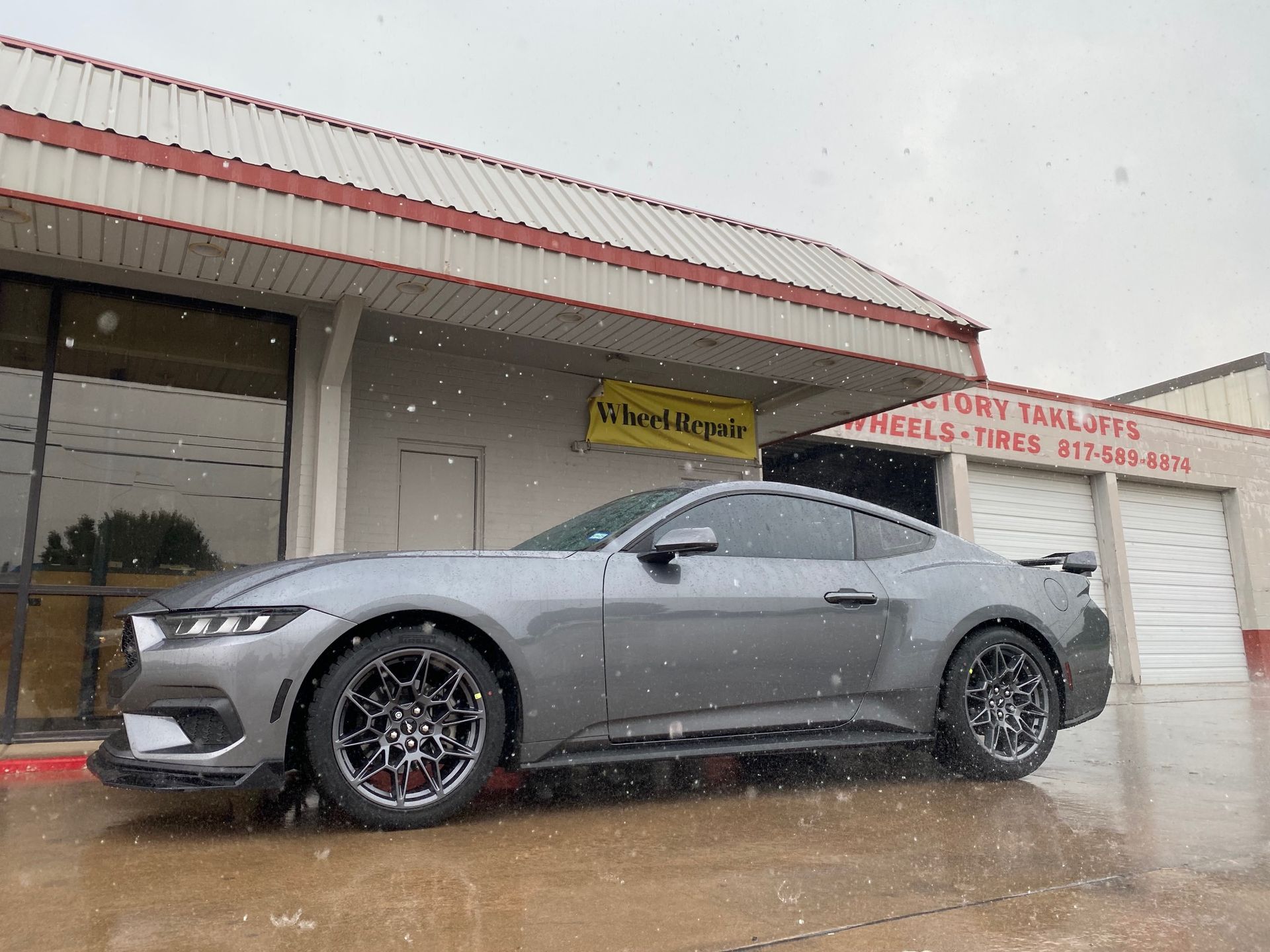 A gray mustang is parked in front of a building in the rain.