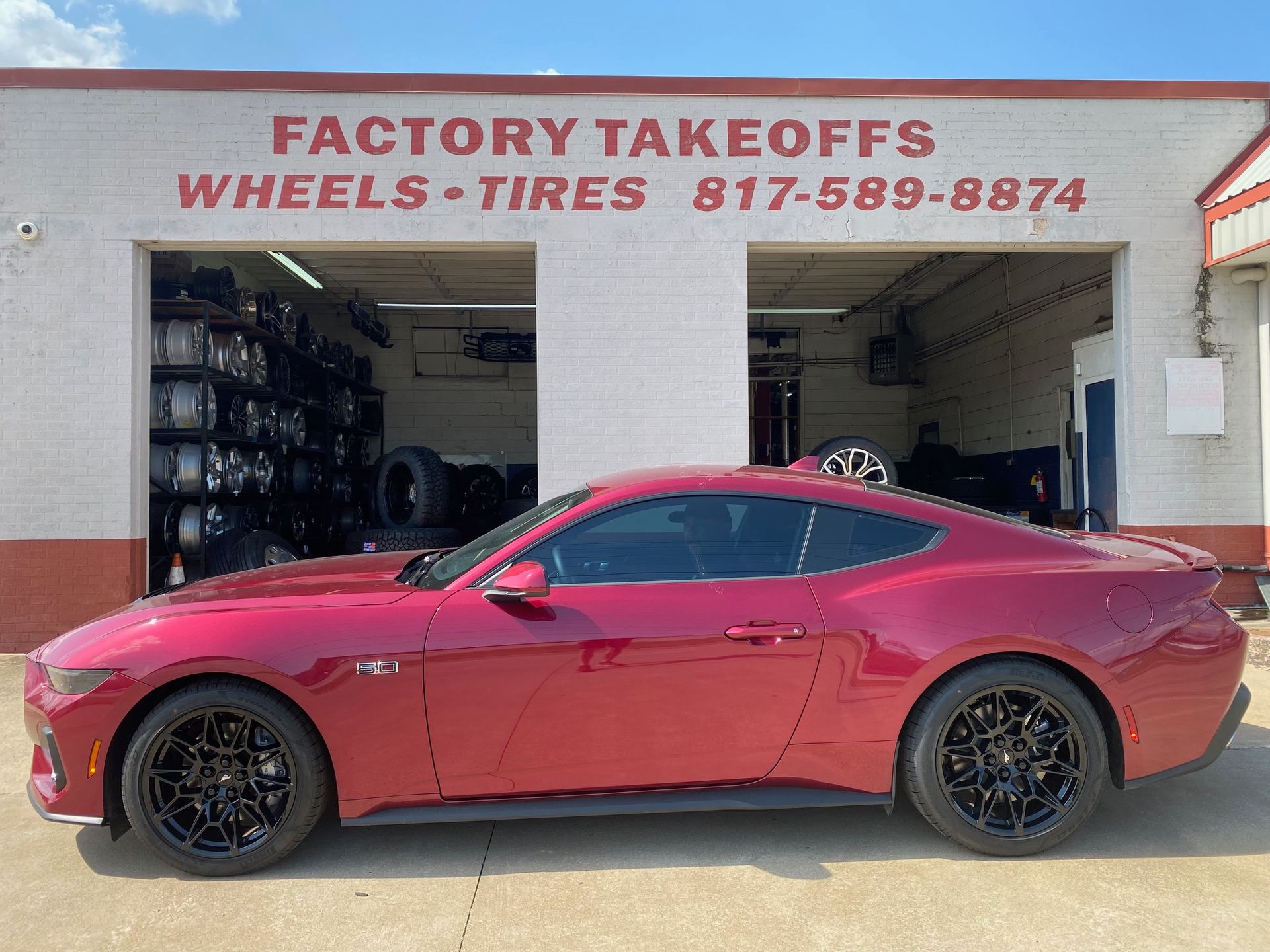 A red mustang is parked in front of a factory takeoffs wheels and tires store.
