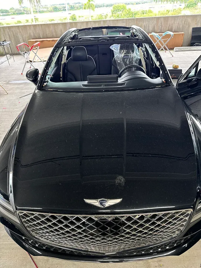 Black luxury car with broken windshield and sunroof, parked outdoors under a covered area.