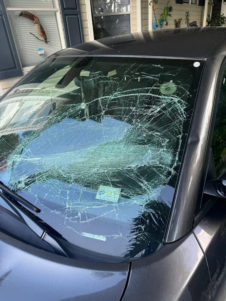 A car windshield completely shattered, displaying a complex spiderweb of cracks. The car is dark gray and parked outside.