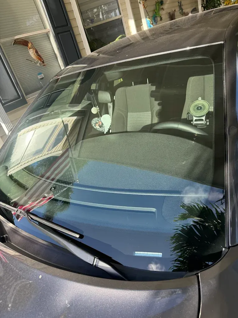 Gray car windshield, interior visible with decorations hanging from the rearview mirror, outdoor setting.