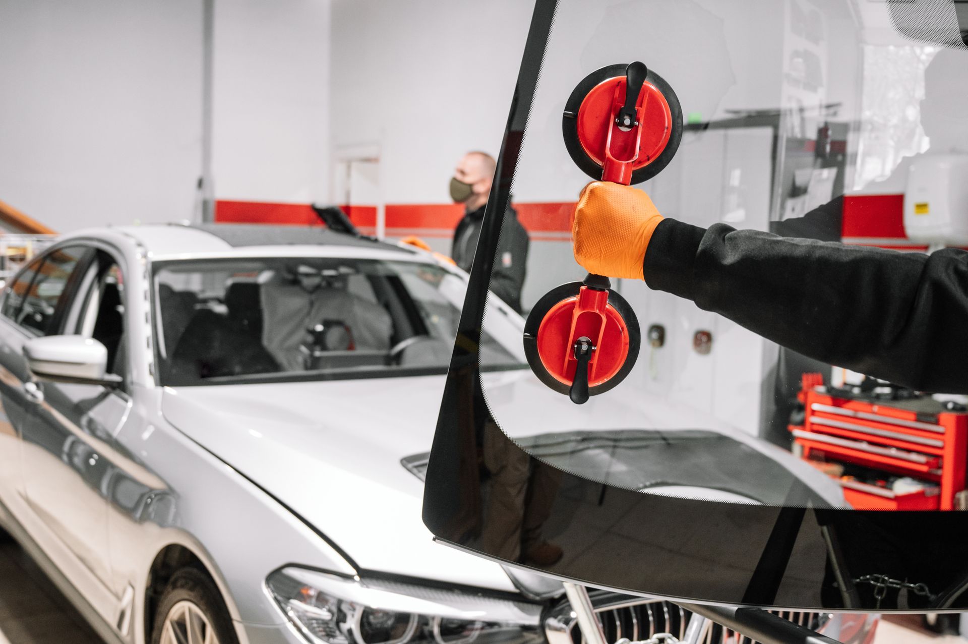A person with orange gloves using suction cups to lift a windshield in a car repair shop. Another person with a face mask is in the background.
