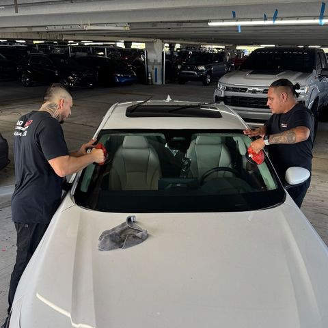 Two men cleaning the windshield of a white car in a parking garage. They are holding red cloths and appear to be working.