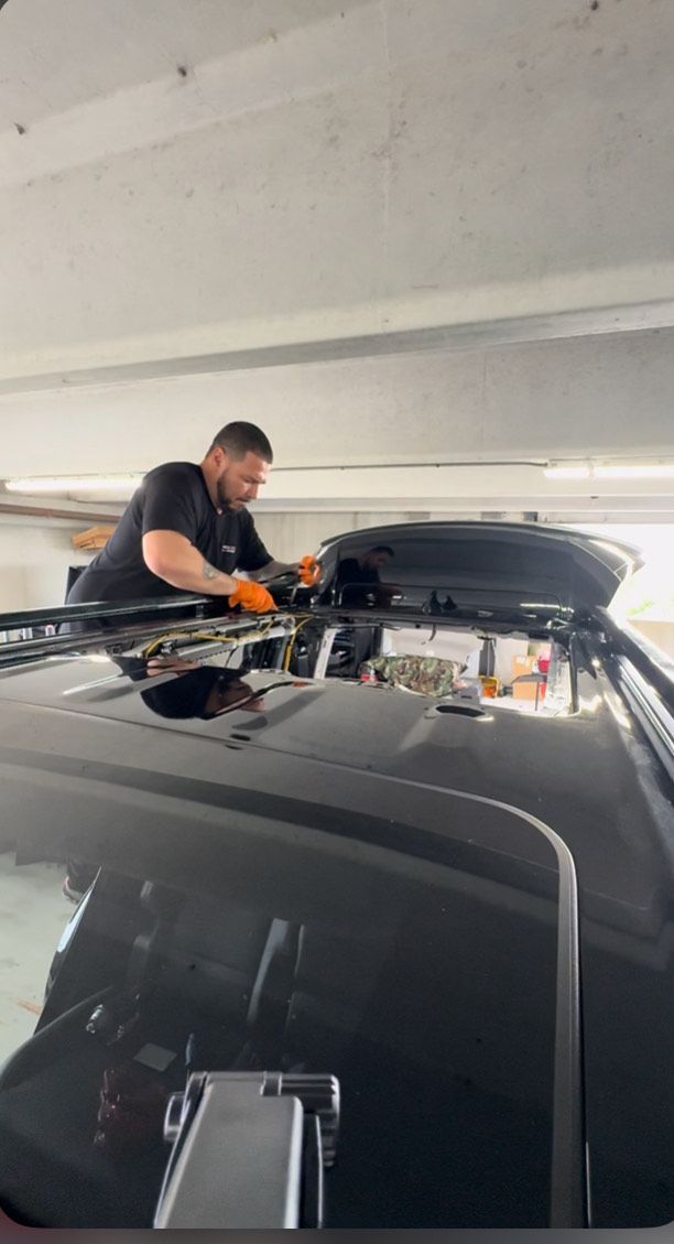 A man is installing a black rooftop cargo box on a black car in a garage. He is wearing gloves and working on the roof.