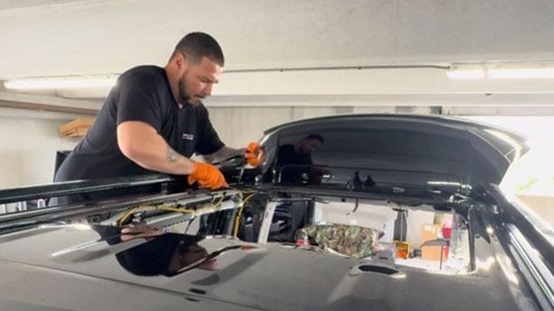 A man in orange gloves works on the roof of a black car, likely installing equipment in a garage.