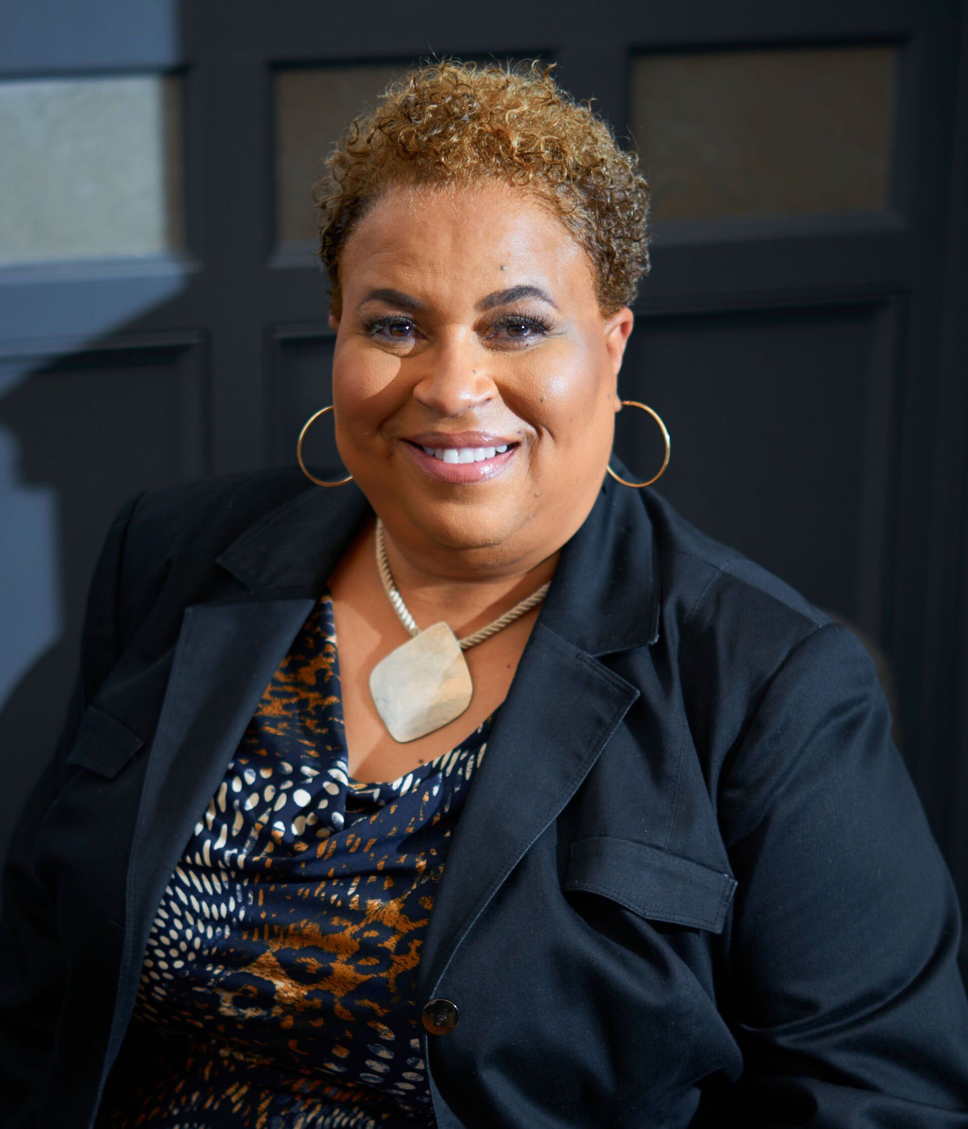 A smiling person wears a black blazer, a patterned top, and a large, light-colored diamond-shaped pendant necklace.