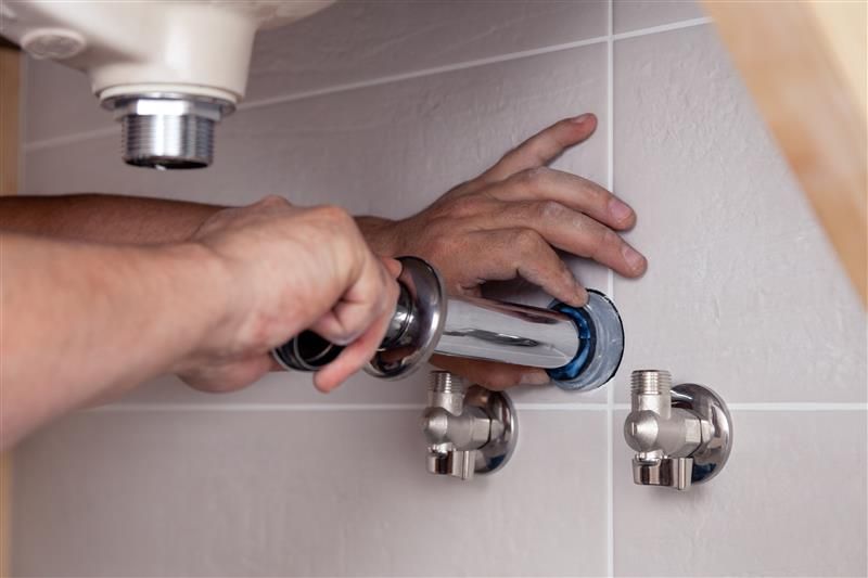 Hands of a plumber installing a chrome pipe under a bathroom sink. Gray tiles and fixtures visible.