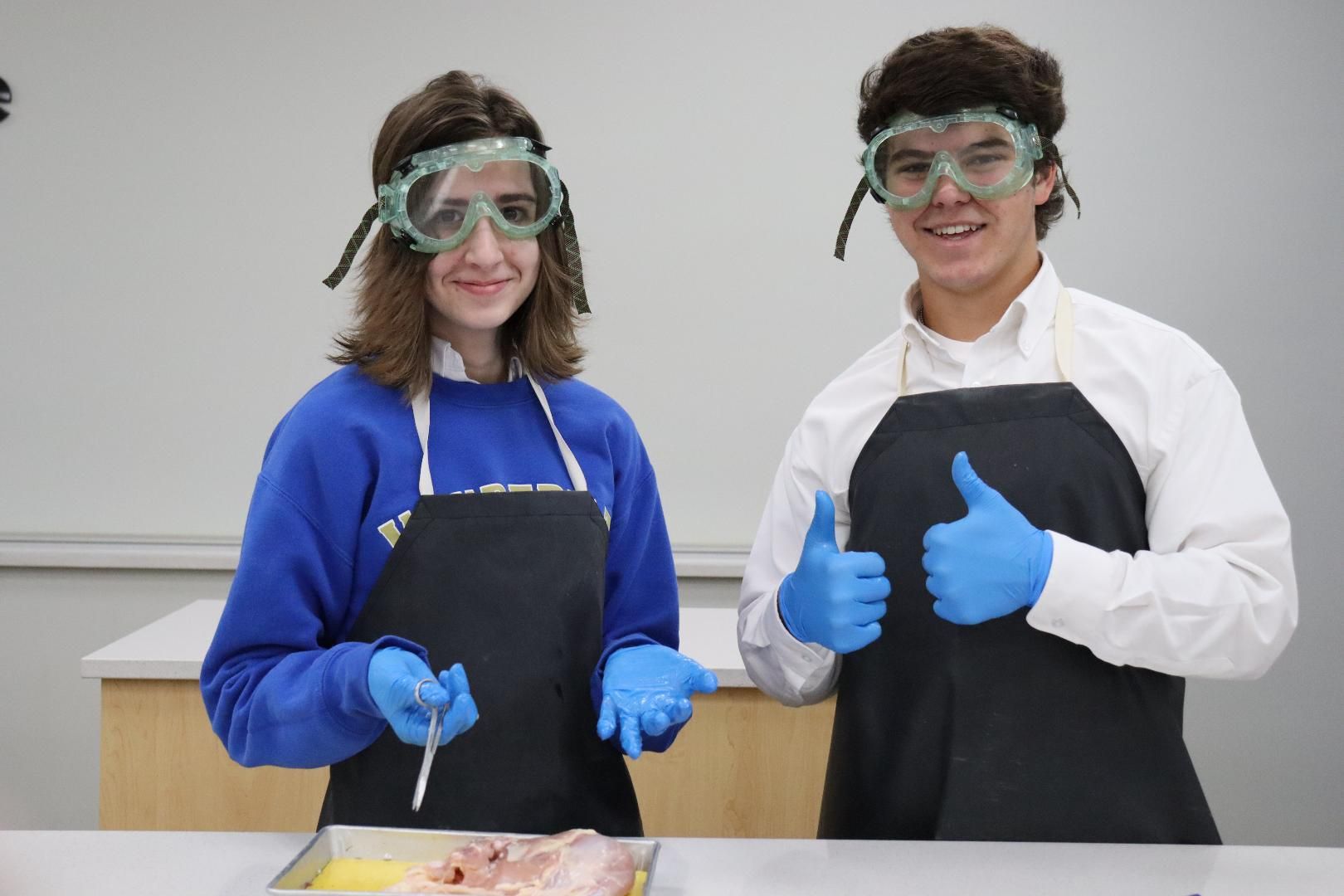 A boy and a girl wearing aprons and goggles are giving a thumbs up