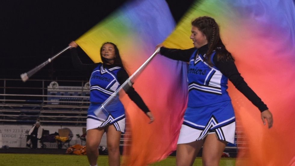 Two cheerleaders are holding rainbow colored flags on a field.