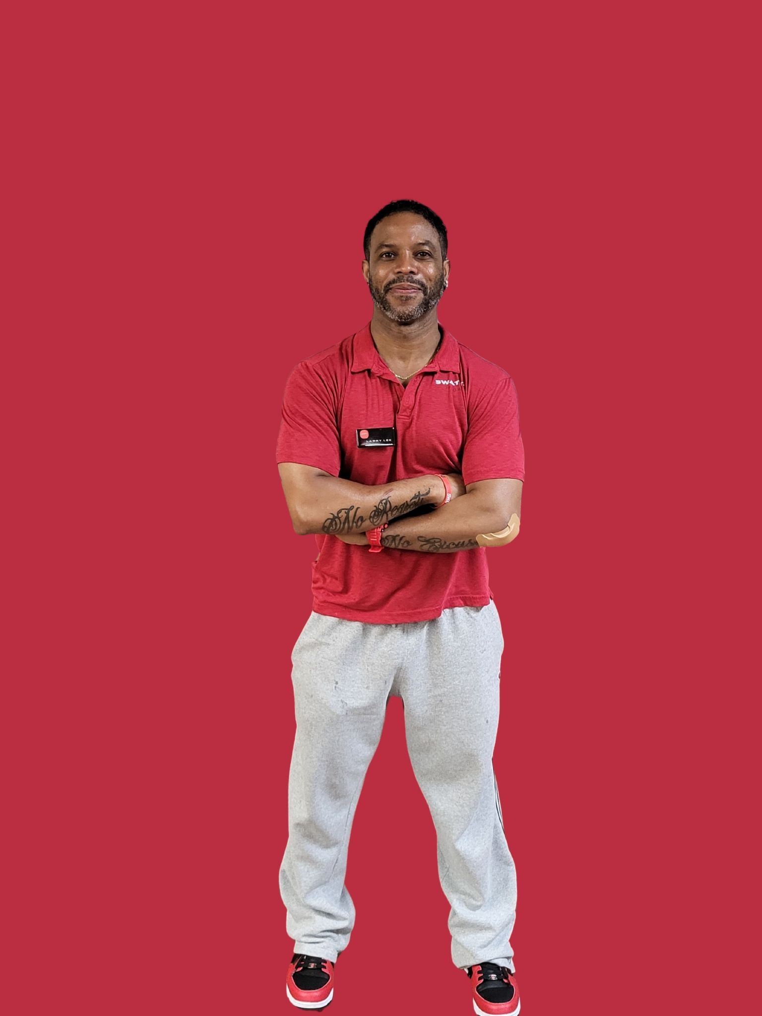 Man in red shirt and gray pants with arms crossed, standing against a red background.