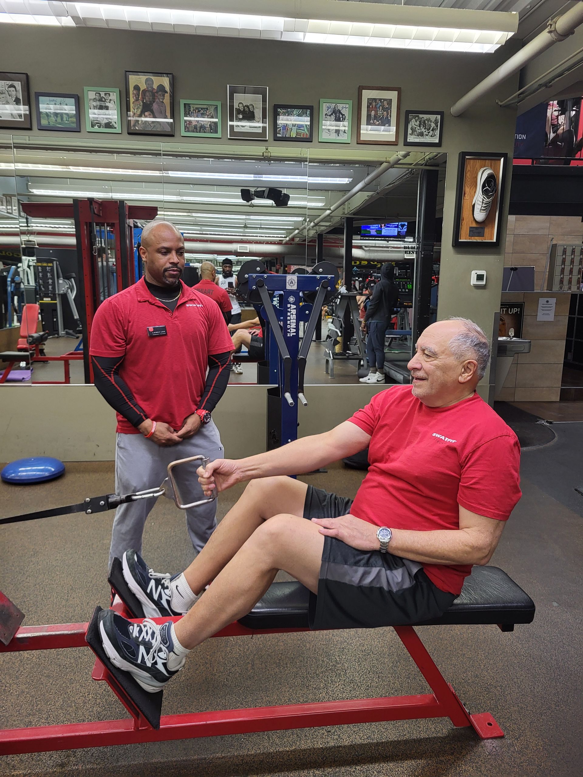Man on weight machine with trainer in gym. Both wear red shirts.